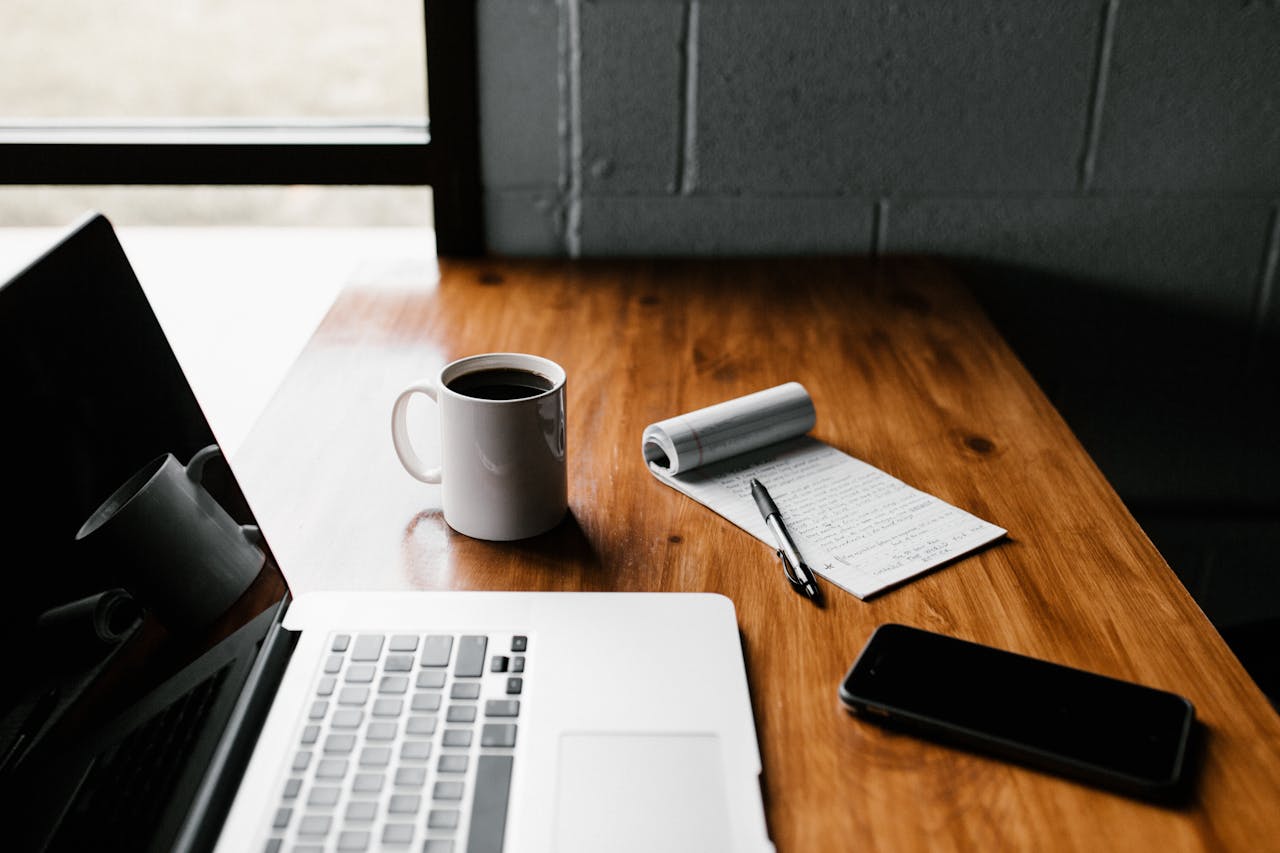 About A tidy workspace featuring a laptop, coffee cup, phone, and notepad on a wooden desk.