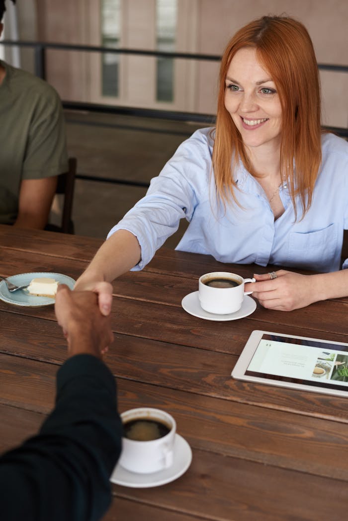 Home Smiling woman with red hair shakes hands across a table during a coffee meeting.