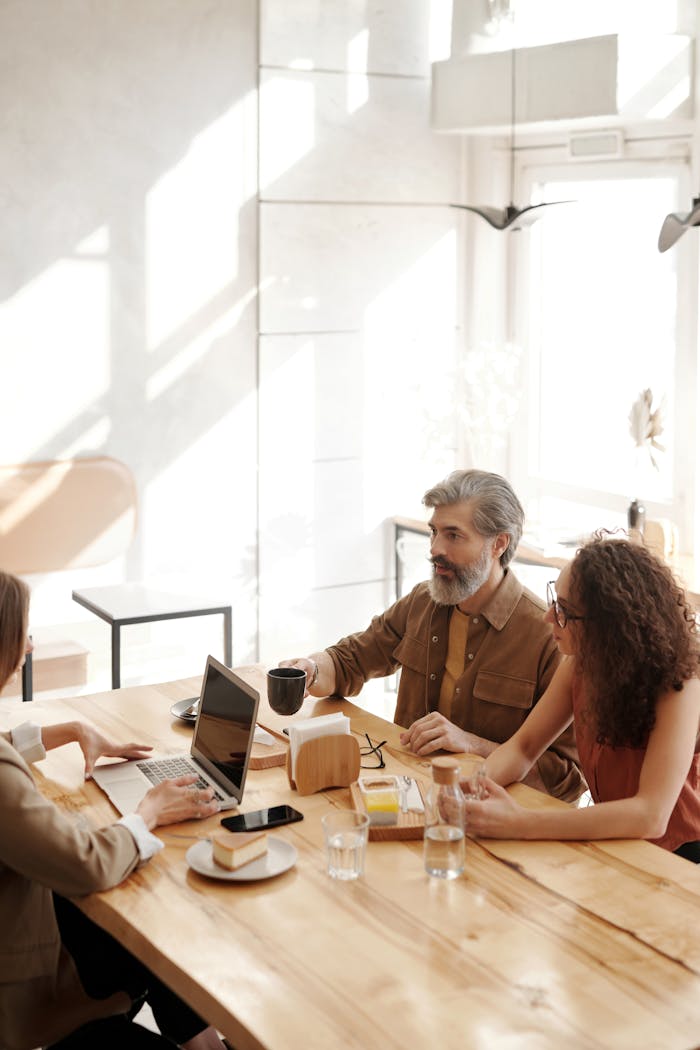 Home Three colleagues in a coffee shop discussing work at a table with laptops and drinks.